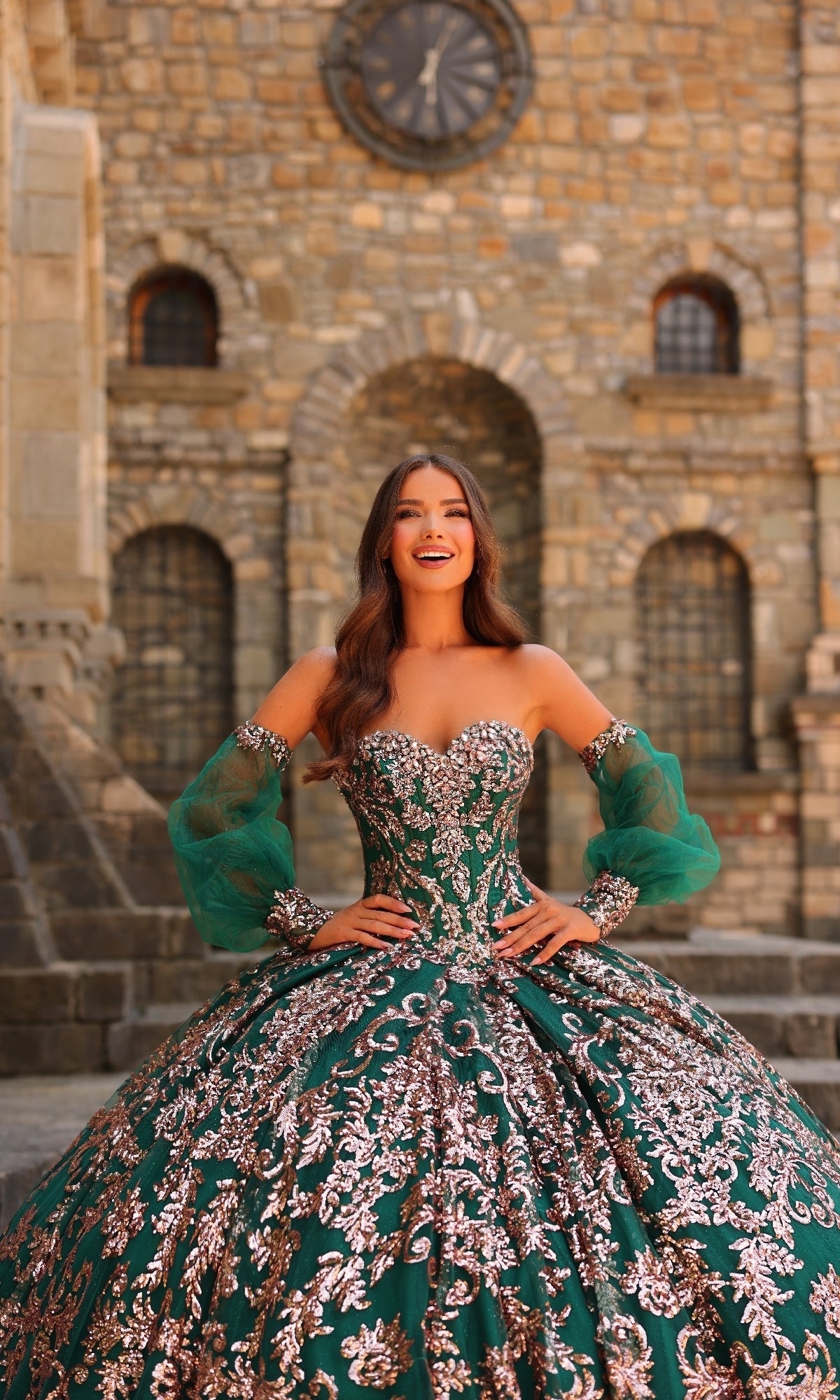 A woman wearing the Amarra 54238 Sequin Quinceanera Dress with puff sleeves smiles in front of a stone building featuring arched windows and a large clock.